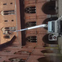 Prace wysokościowe przy ceglanej fasadzie zamku z użyciem podnośnika koszowego, widoczna platforma z pracownikiem na tle gotyckich okien.
