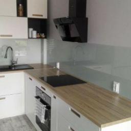 JB-Decor - Nowoczesna, jasna kuchnia z białymi szafkami, drewnianym blatem, czarnym zlewem i płytą indukcyjną oraz szklanym panelem ściennym w odcieniach szarości.