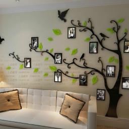 JB-Decor - Ściana salonu z dekoracją w postaci czarnego drzewa z ramkami na zdjęcia i zielonymi liśćmi, nad białą sofą z poduszkami.