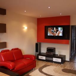 JB-Decor - Jasne wnętrze salonu z czerwoną sofą, telewizorem na czerwonej ścianie akcentującej i drewnianą podłogą z wzorzystym dywanem.