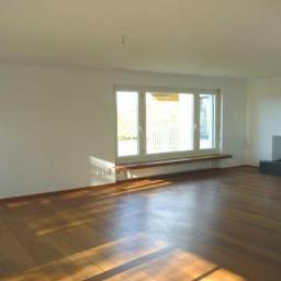 JB-Decor - Pusty pokój z drewnianą podłogą, dużym oknem z parapetem i nowoczesnym kominkiem w rogu. Ściany świeżo pomalowane na biało, brak mebli.