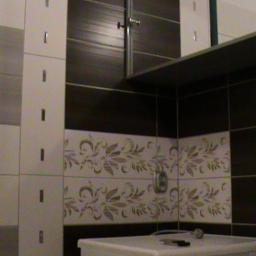 JB-Decor - Wykończona łazienka z brązowo-białymi płytkami, dekoracyjnym motywem roślinnym na ścianie, białą szafką z lustrem i półką, widoczne akcesoria łazienkowe na umywalce.