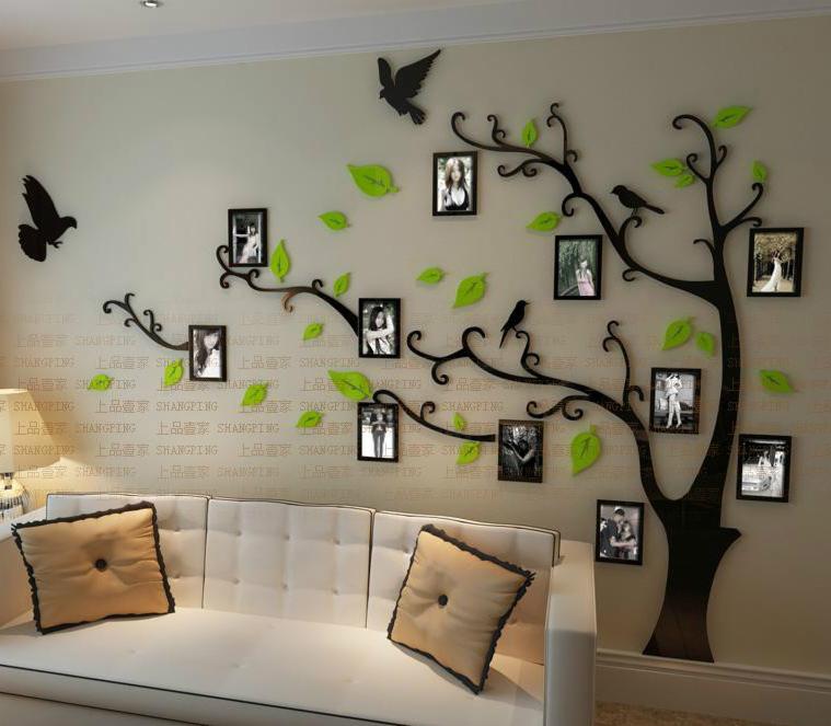 Ściana salonu z dekoracją w postaci czarnego drzewa z ramkami na zdjęcia i zielonymi liśćmi, nad białą sofą z poduszkami.
