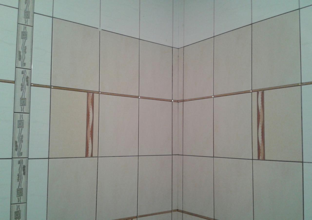 Wykończony narożnik łazienki z beżowymi i białymi płytkami, ozdobny pas z motywem geometrycznym po lewej stronie.