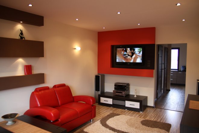 Jasne wnętrze salonu z czerwoną sofą, telewizorem na czerwonej ścianie akcentującej i drewnianą podłogą z wzorzystym dywanem.