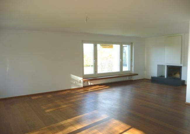 Pusty pokój z drewnianą podłogą, dużym oknem z parapetem i nowoczesnym kominkiem w rogu. Ściany świeżo pomalowane na biało, brak mebli.