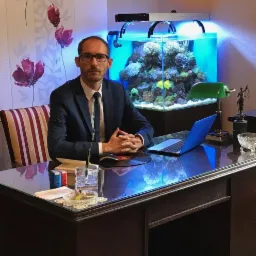 Mężczyzna w garniturze siedzi za biurkiem z akwarium w tle, na biurku laptop, lampka i statuetka Temidy.