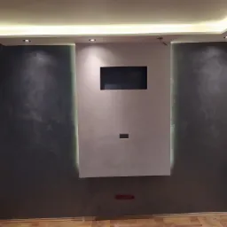 Ściana z dekoracyjną szarą fakturą i podświetlanym panelem w kolorze jasnego beżu z wnękami na TV i gniazdka, parkiet w jodełkę, oświetlenie sufitowe LED.