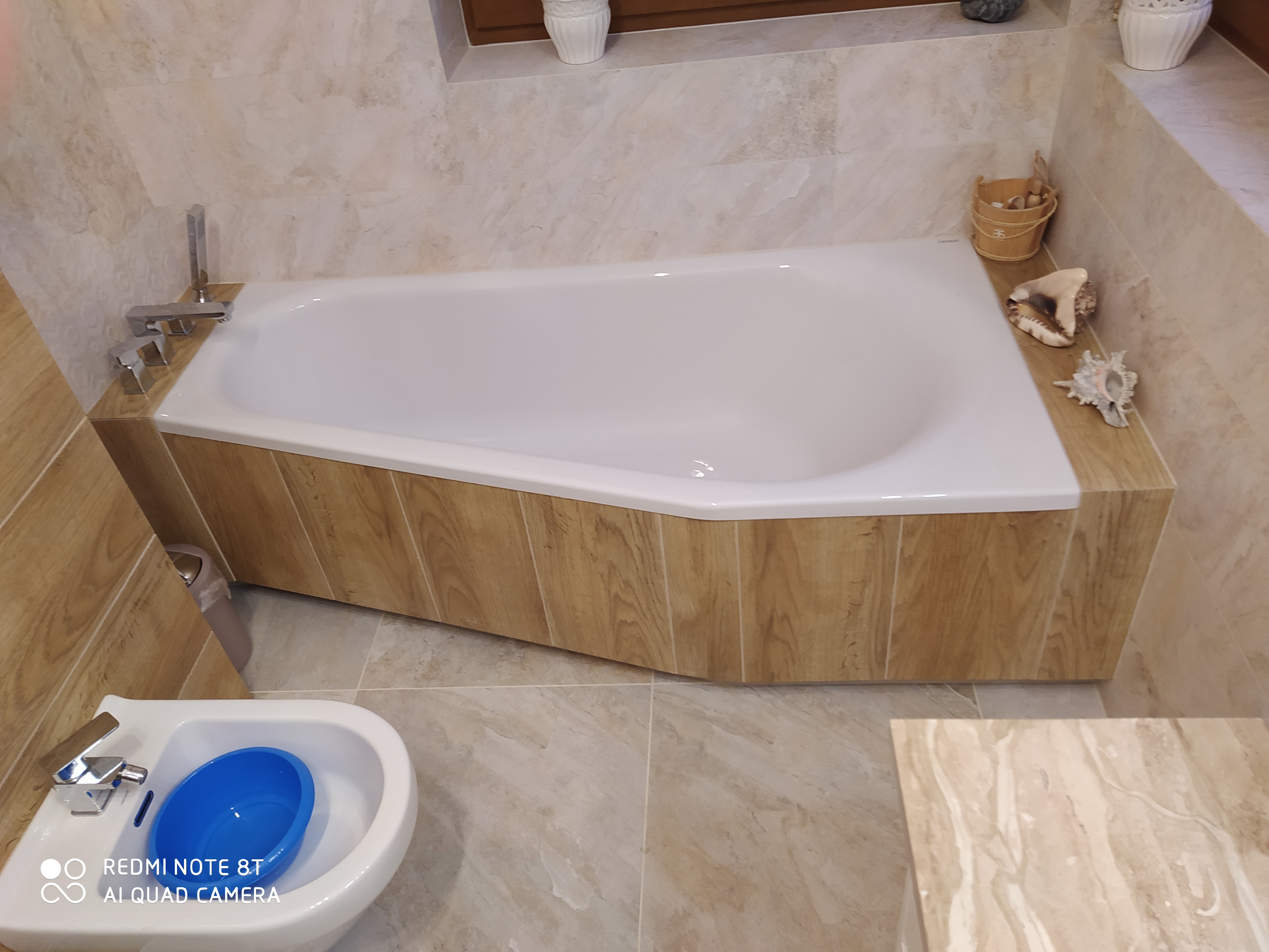 Wykończona łazienka z wanną obudowaną panelami imitującymi drewno, bidetem z niebieską miską i dekoracjami z muszli.