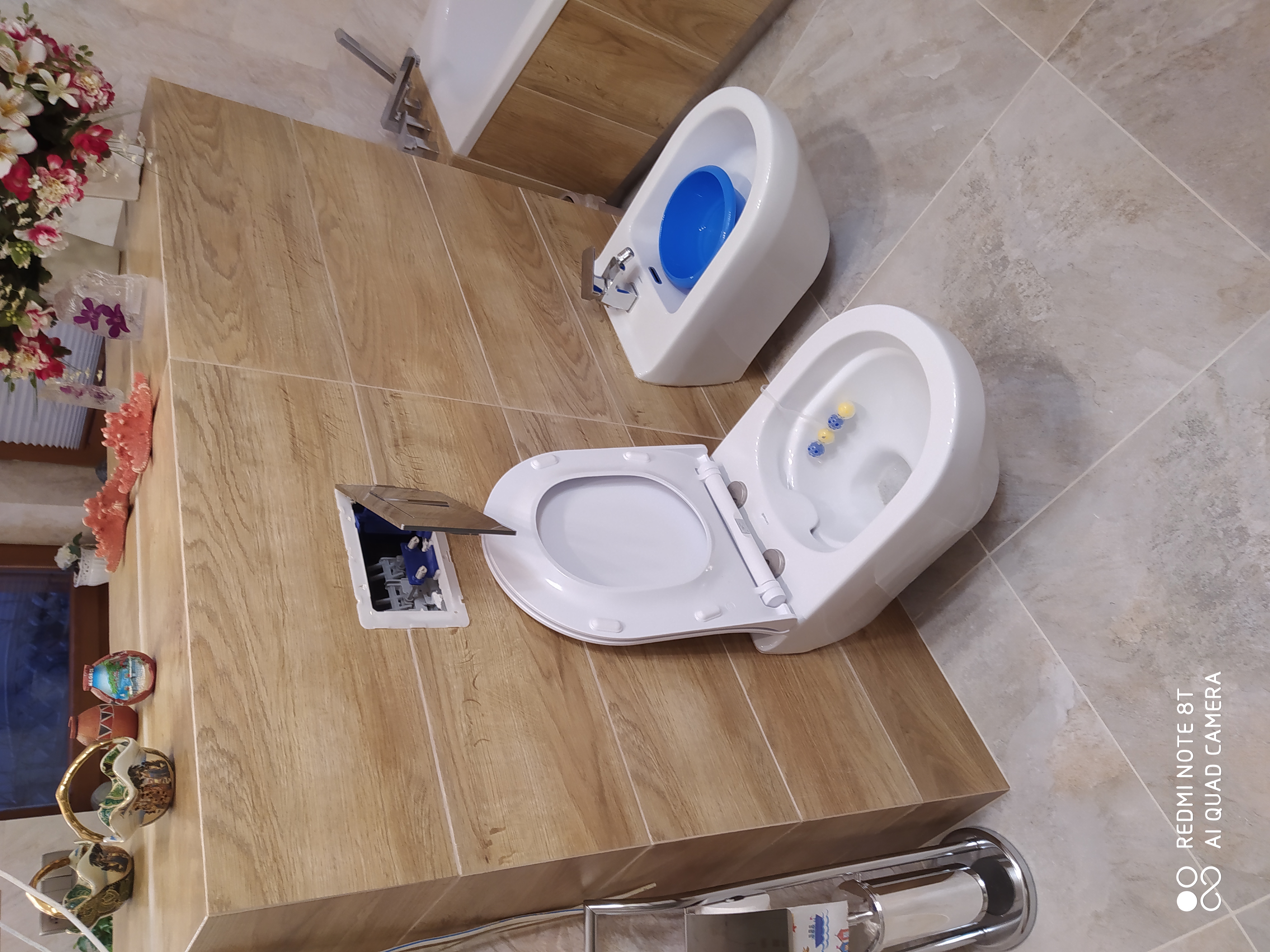 Biała toaleta i bidet zamontowane na płytkach imitujących drewno, z otwartym włazem rewizyjnym na ścianie.