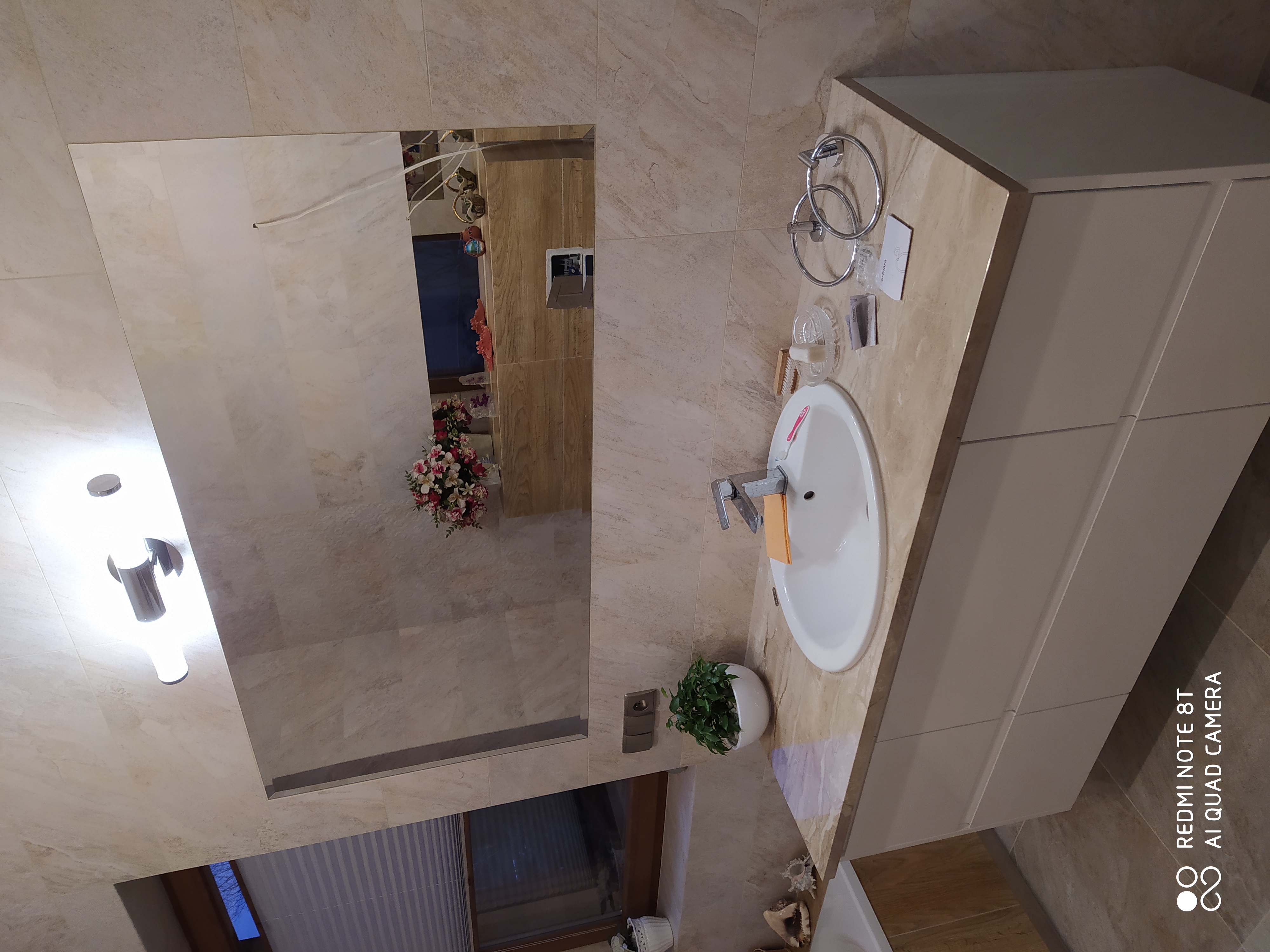 Umywalka nablatowa z szarą szafką w łazience wykończonej beżowymi płytkami, widok z lustra na kwiaty w pokoju obok.