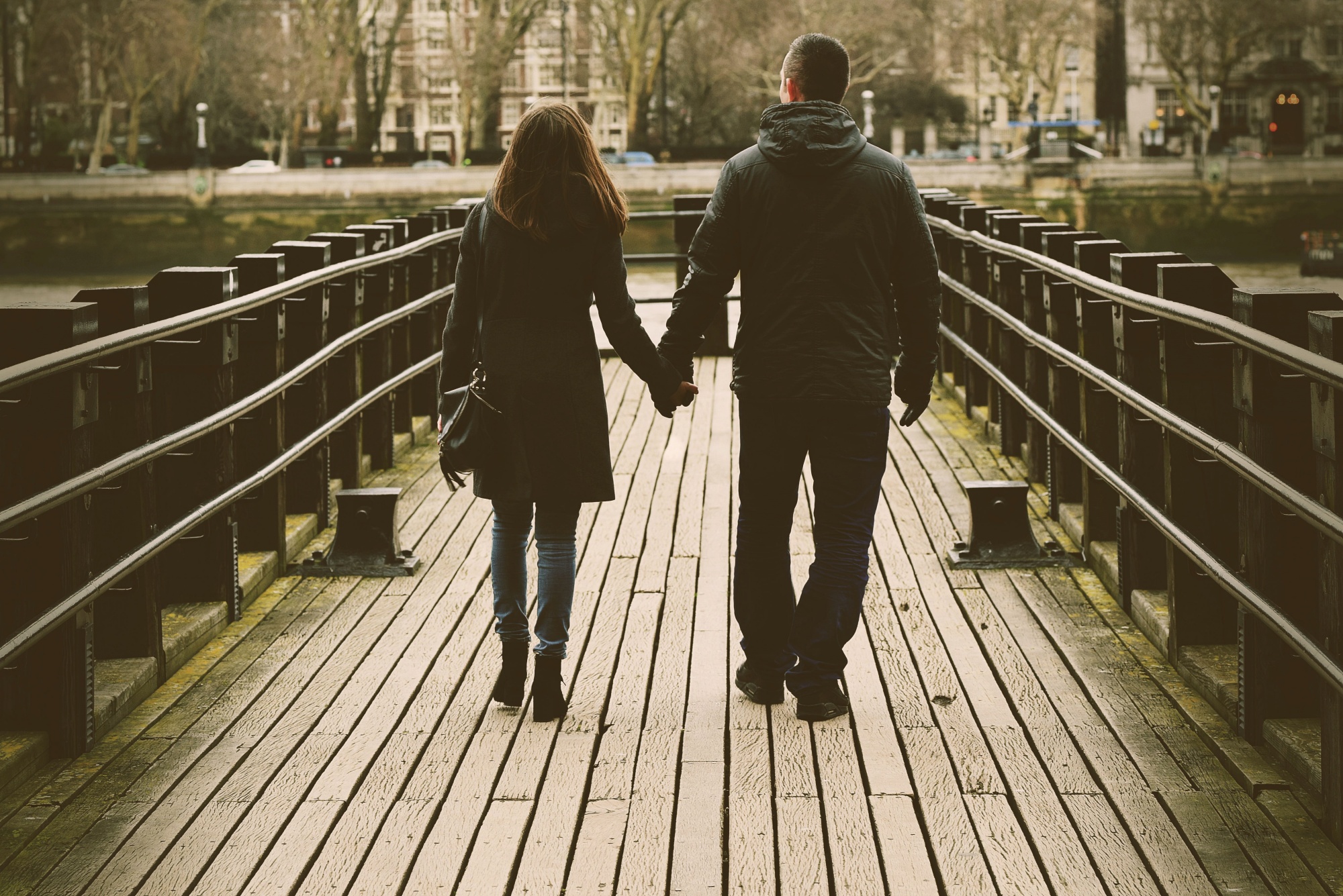 Para trzymająca się za ręce, spacerująca po drewnianym pomoście w jesiennej scenerii miejskiej, widok z tyłu.