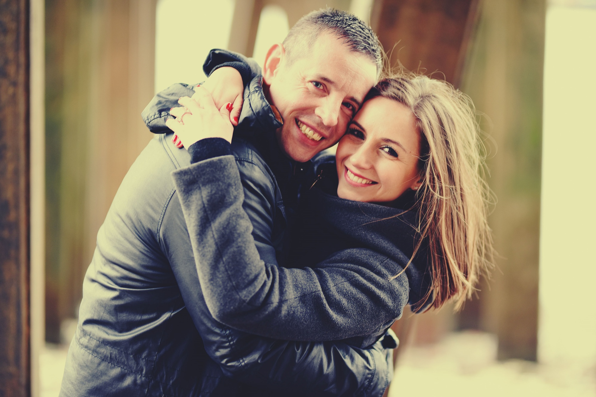 Uśmiechnięta para, mężczyzna w skórzanej kurtce obejmuje kobietę w szarym płaszczu, na palcu kobiety widoczny pierścionek zaręczynowy, ciepłe oświetlenie.