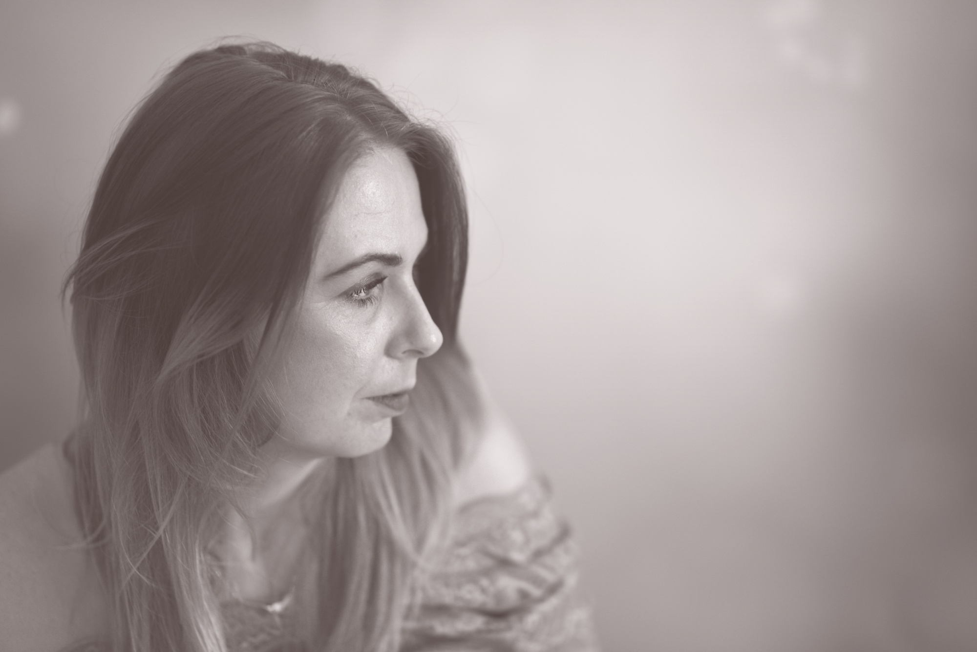 Portret kobiety z profilu, z długimi włosami, skierowanej w lewo, na rozmytym tle w odcieniach szarości.