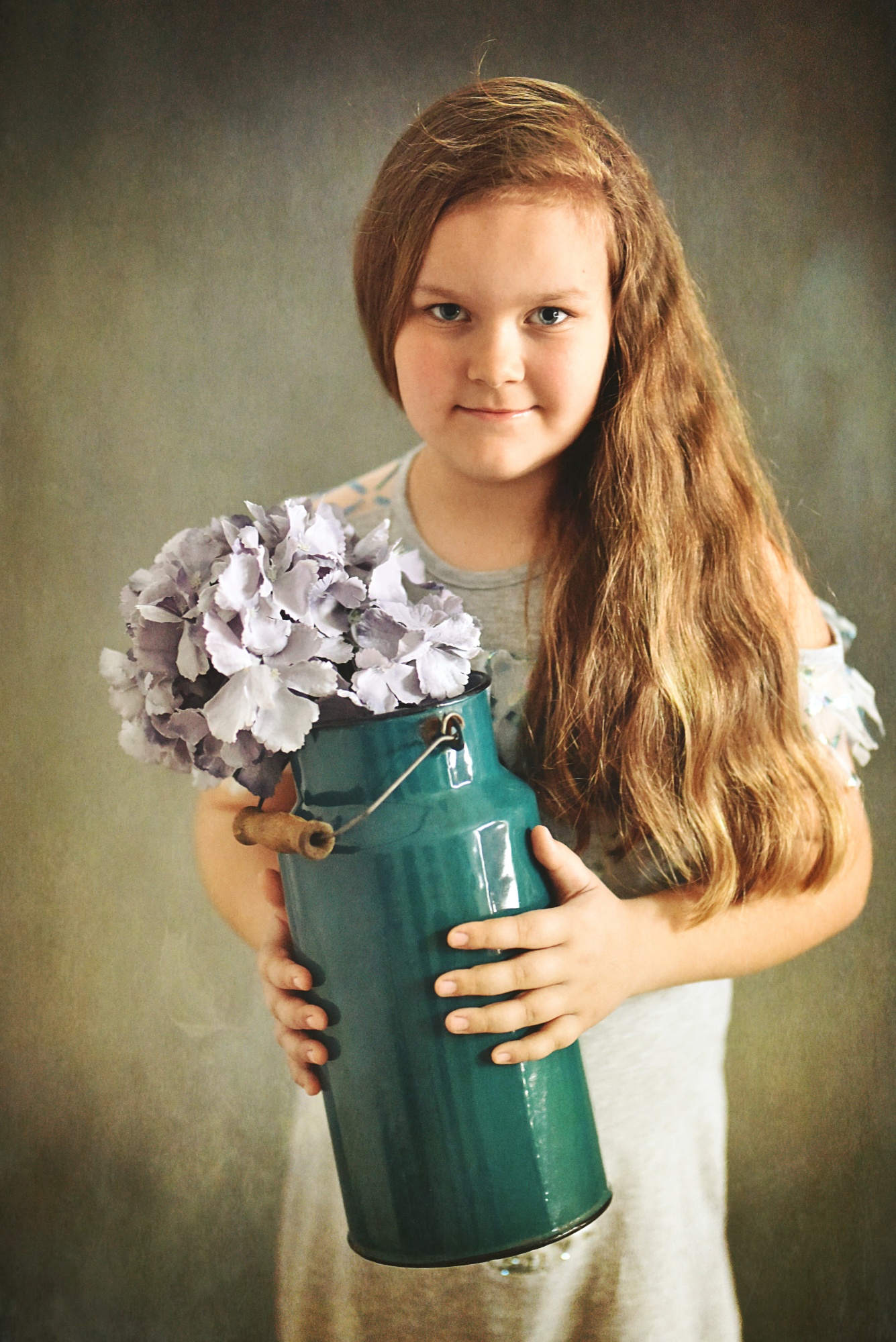 Dziewczynka z długimi włosami trzyma turkusowy, emaliowany dzbanek z bukietem fioletowych hortensji na rozmytym szarym tle.