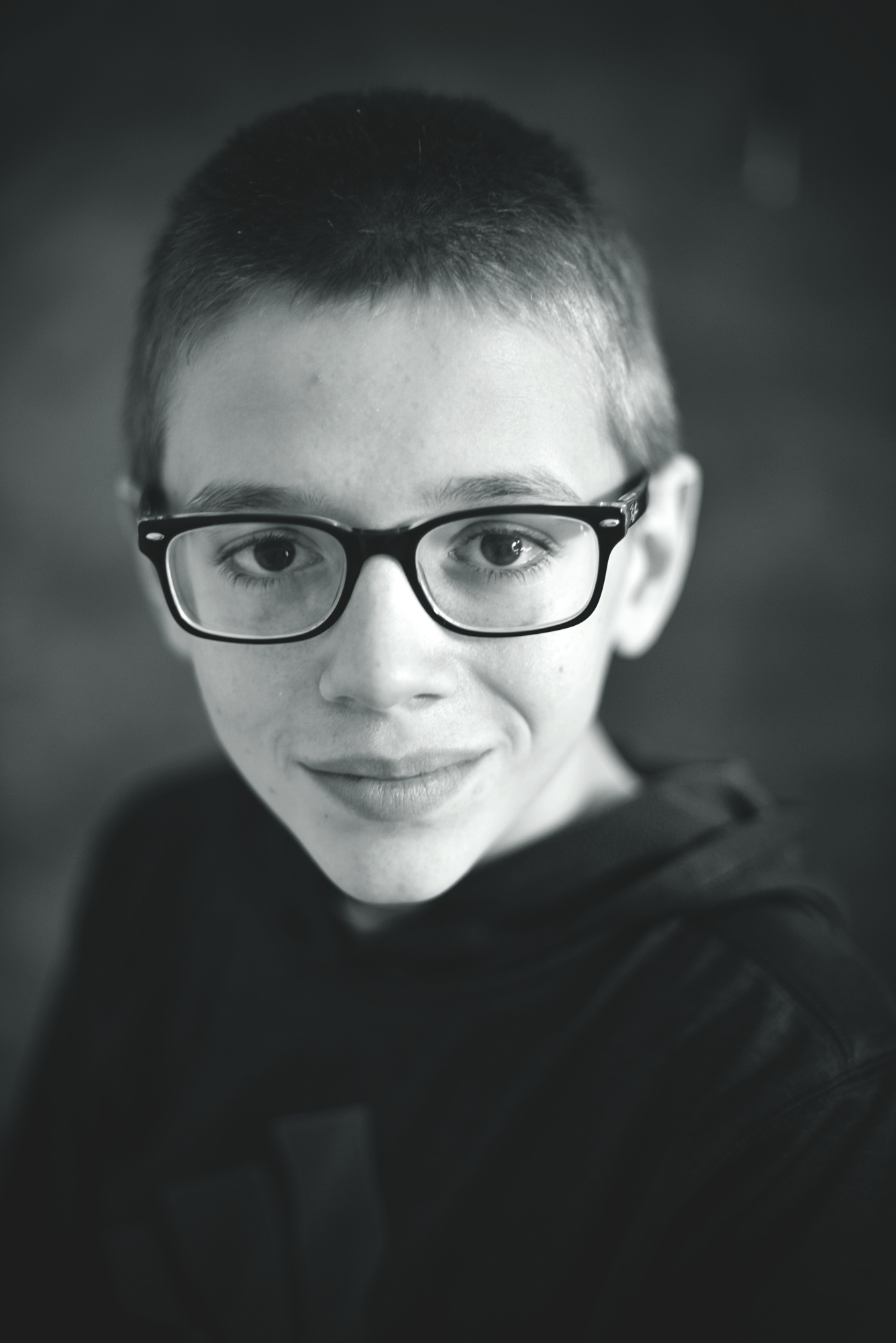 Portret czarno-biały uśmiechniętego nastolatka w okularach z ciemnymi oprawkami, z krótkimi włosami i w ciemnej bluzie z kapturem, wykonany z bliska.