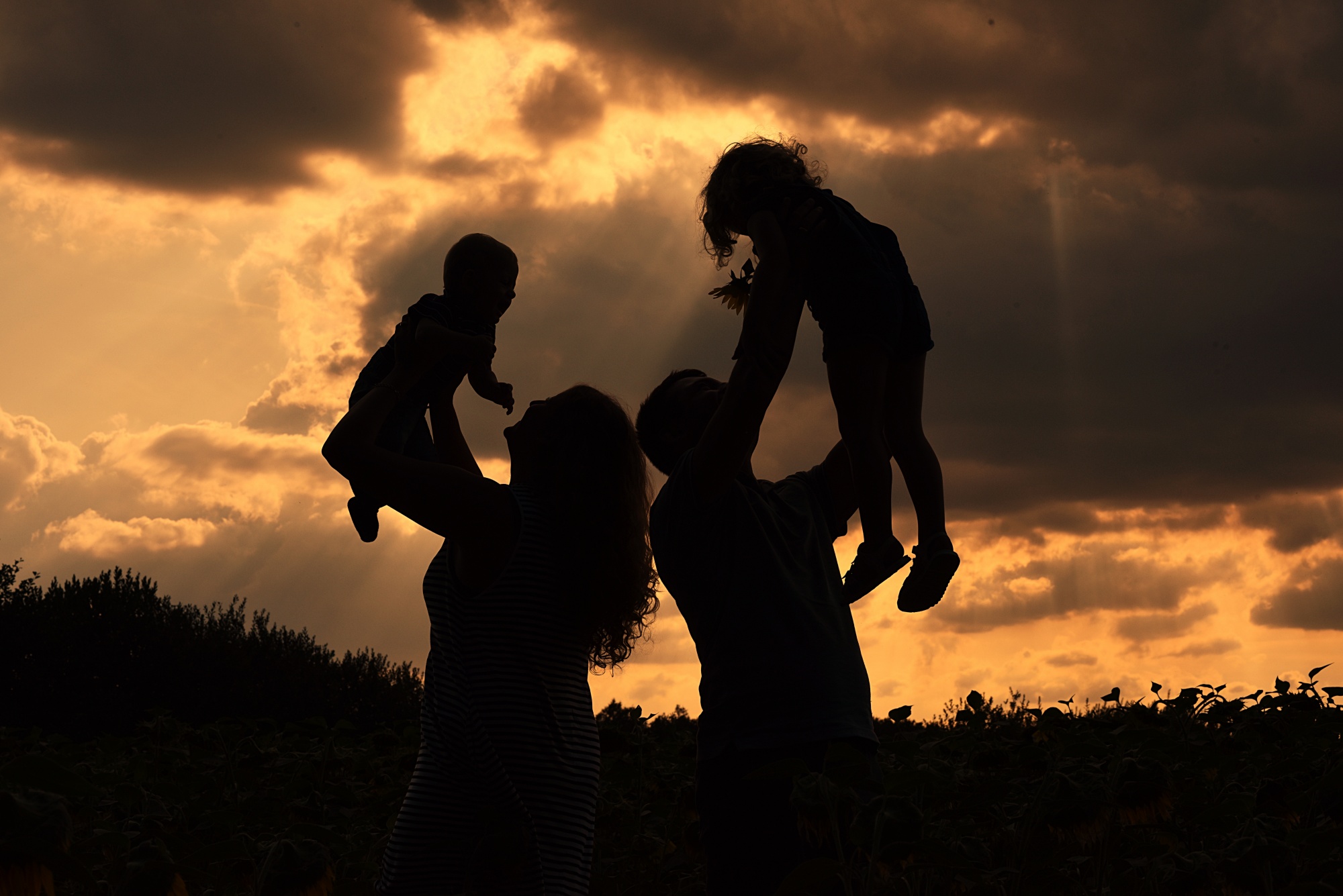 Silhouettes of a family with two children against an orange sunset sky in Jelcz-Laskowice, one parent holding a baby, the other holding a toddler with a flower.