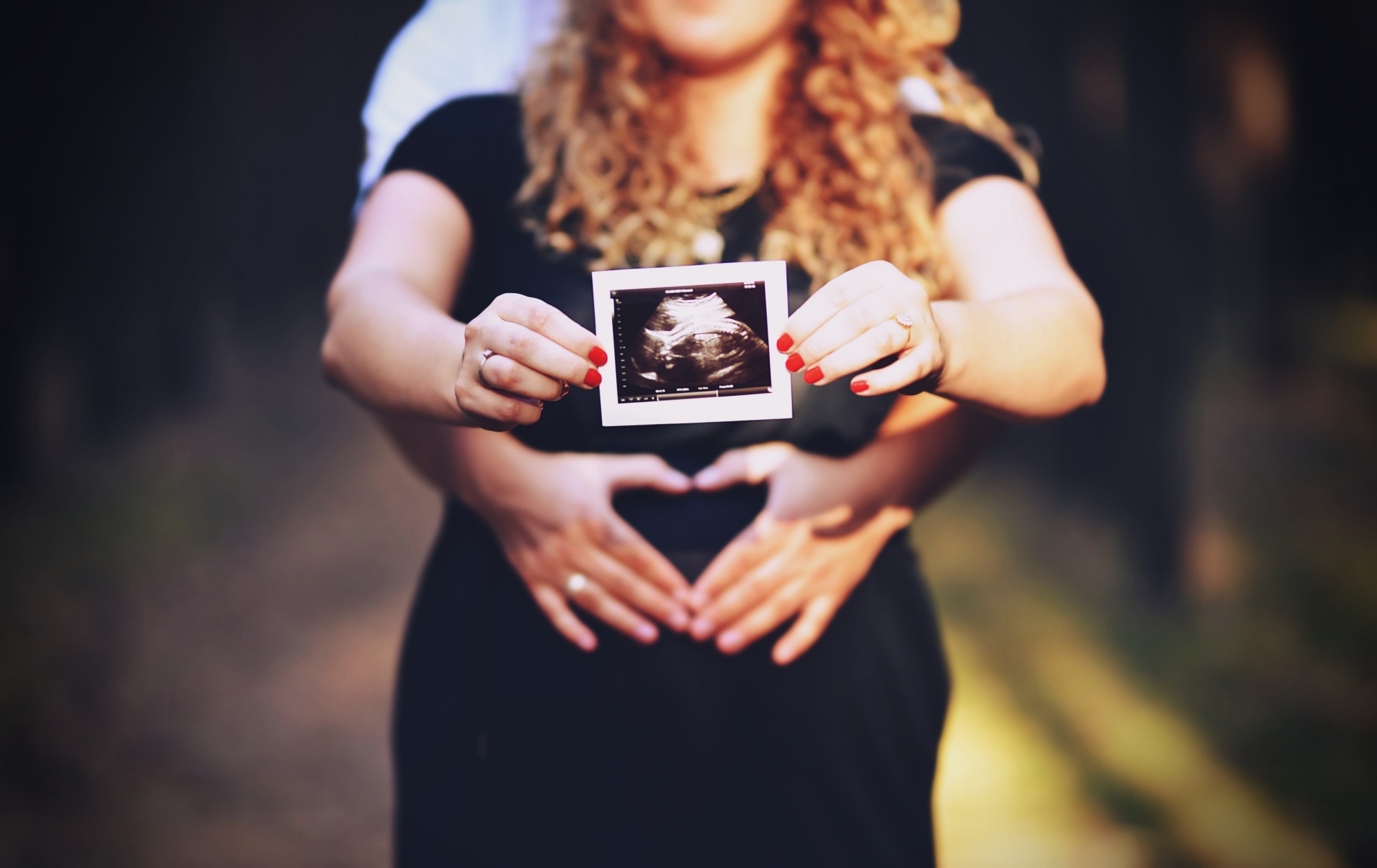 Przyszła mama w czarnej sukience trzyma zdjęcie USG, a dłonie partnera obejmują ją, tworząc kształt serca na brzuchu.