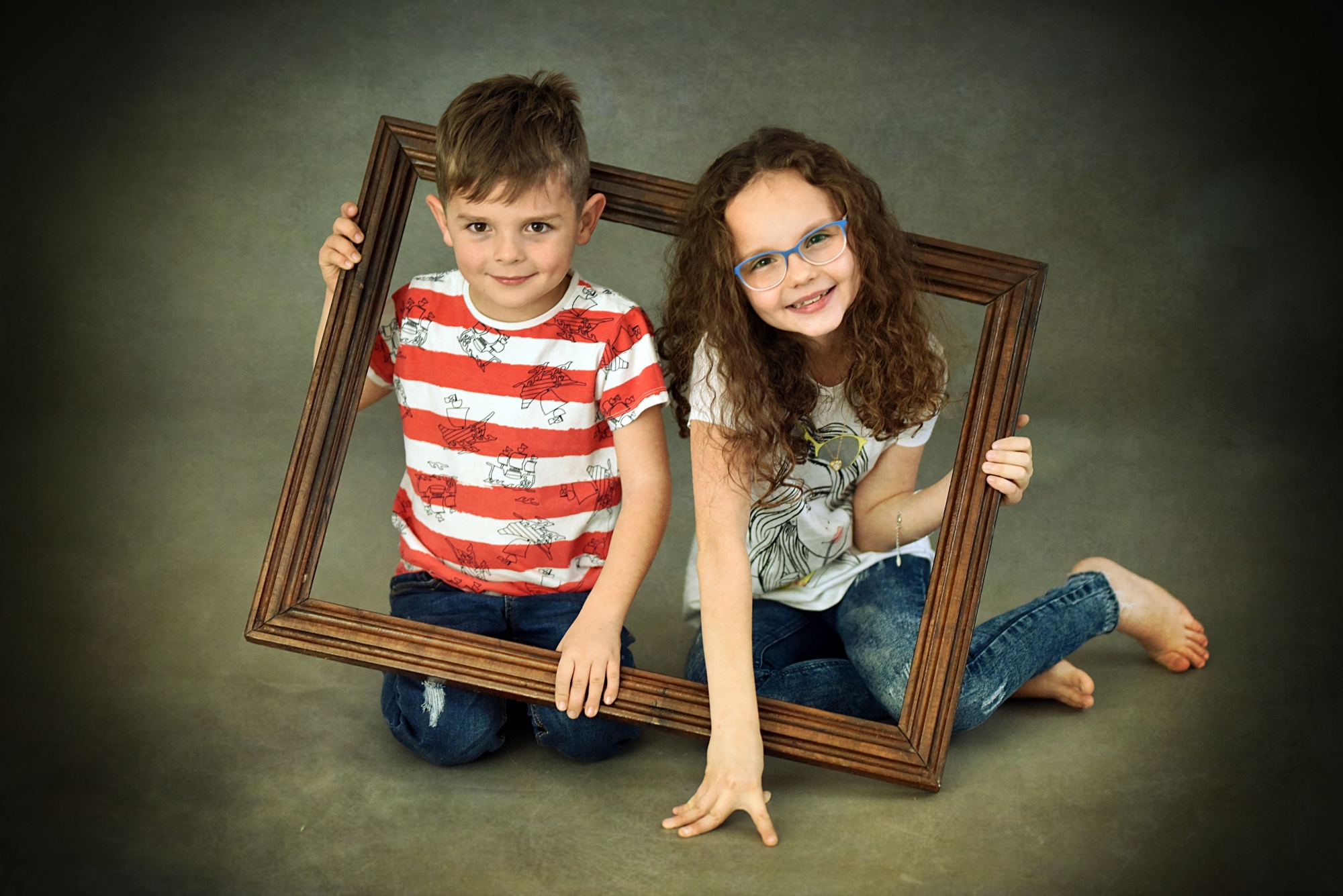 Dwoje uśmiechniętych dzieci, chłopiec w koszulce w paski i dziewczynka w okularach, pozują z drewnianą ramą na szarym tle.