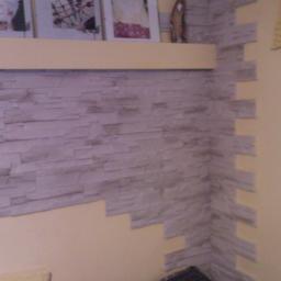 Wadowski Łukasz - Wykończona ściana z dekoracyjnej imitacji kamienia w odcieniach szarości, ułożona do połowy wysokości ściany, z półką i ramkami zdjęć powyżej, oraz żółtym zegarem na sąsiedniej ścianie.