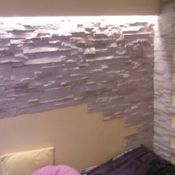 Wadowski Łukasz - Ściana wykończona dekoracyjną okładziną z szarego kamienia imitującego łupek, z podświetleniem LED u góry, częściowo przylegająca do pomalowanej ściany w odcieniu ciepłego beżu, widoczny narożnik...