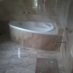 Narożna wanna z hydromasażem obudowana płytkami imitującymi marmur, dopasowane płytki na podłodze i ścianach, widoczne przyłącze wodne.