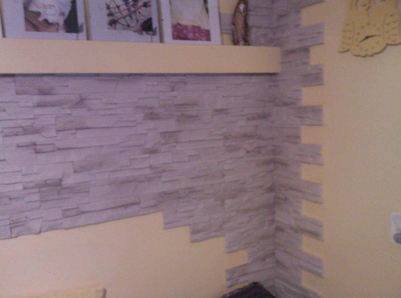 Wykończona ściana z dekoracyjnej imitacji kamienia w odcieniach szarości, ułożona do połowy wysokości ściany, z półką i ramkami zdjęć powyżej, oraz żółtym zegarem na sąsiedniej ścianie.