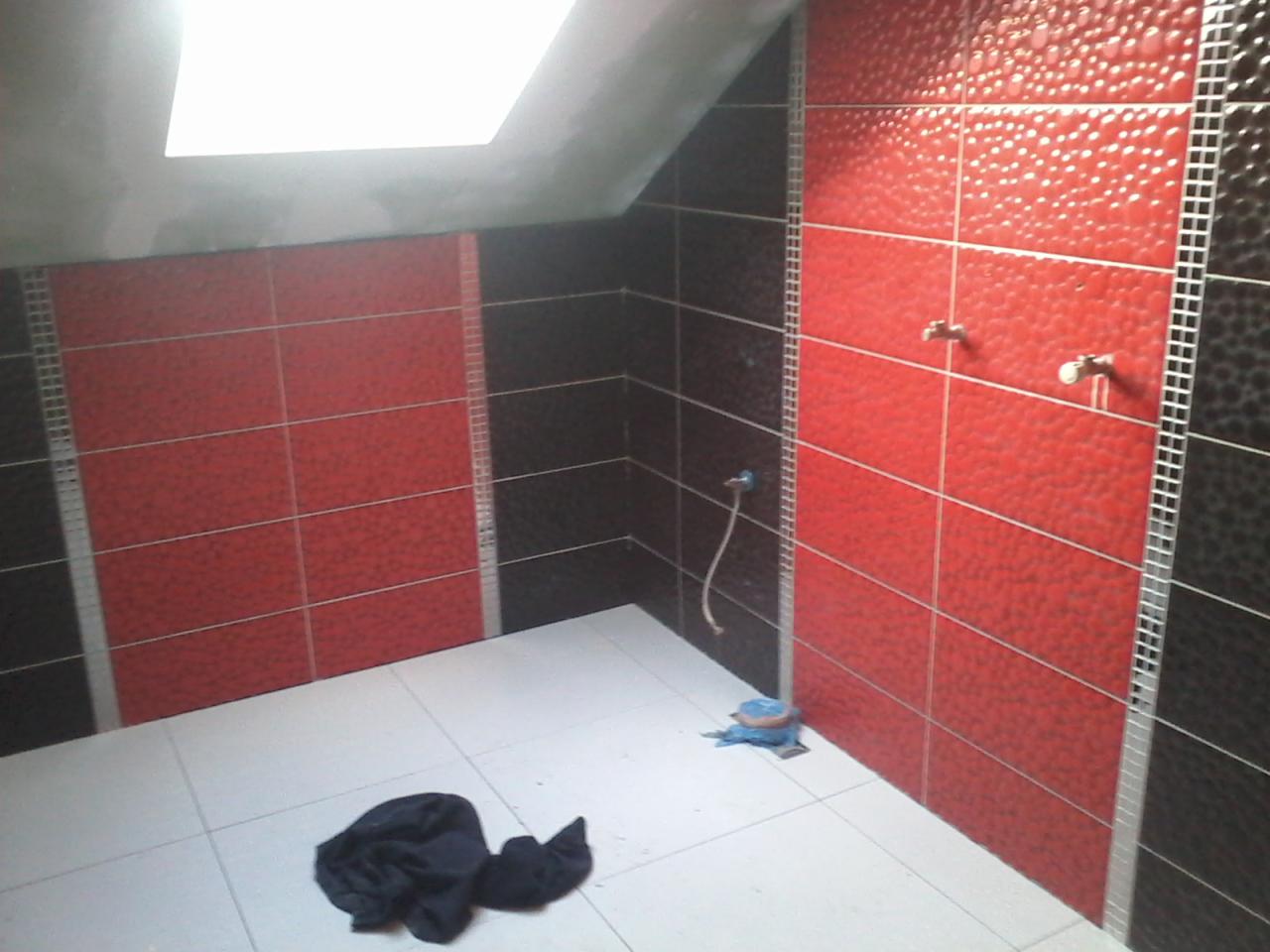 Ściany łazienki wyłożone czerwono-czarnymi płytkami z wyraźną fakturą, widoczne dwa krany, biała podłoga z leżącą szmatką i ubraniem, skośny sufit z oknem.