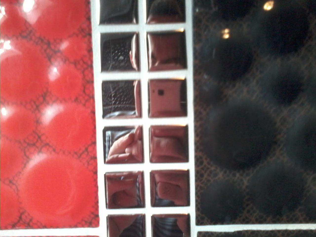 Mozaika ścienna z czerwonymi, czarnymi i lustrzanymi kwadratami, ułożona w rzędach i kolumnach z białymi fugami, ukazująca odbicie dłoni w jednym z lustrzanych elementów.