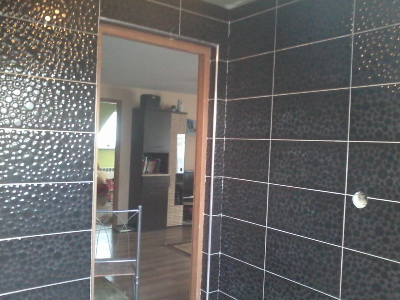 Ciemne, wzorzyste płytki ceramiczne na ścianie i wokół ościeżnicy drzwiowej; widoczny fragment wnętrza z meblami w tle.