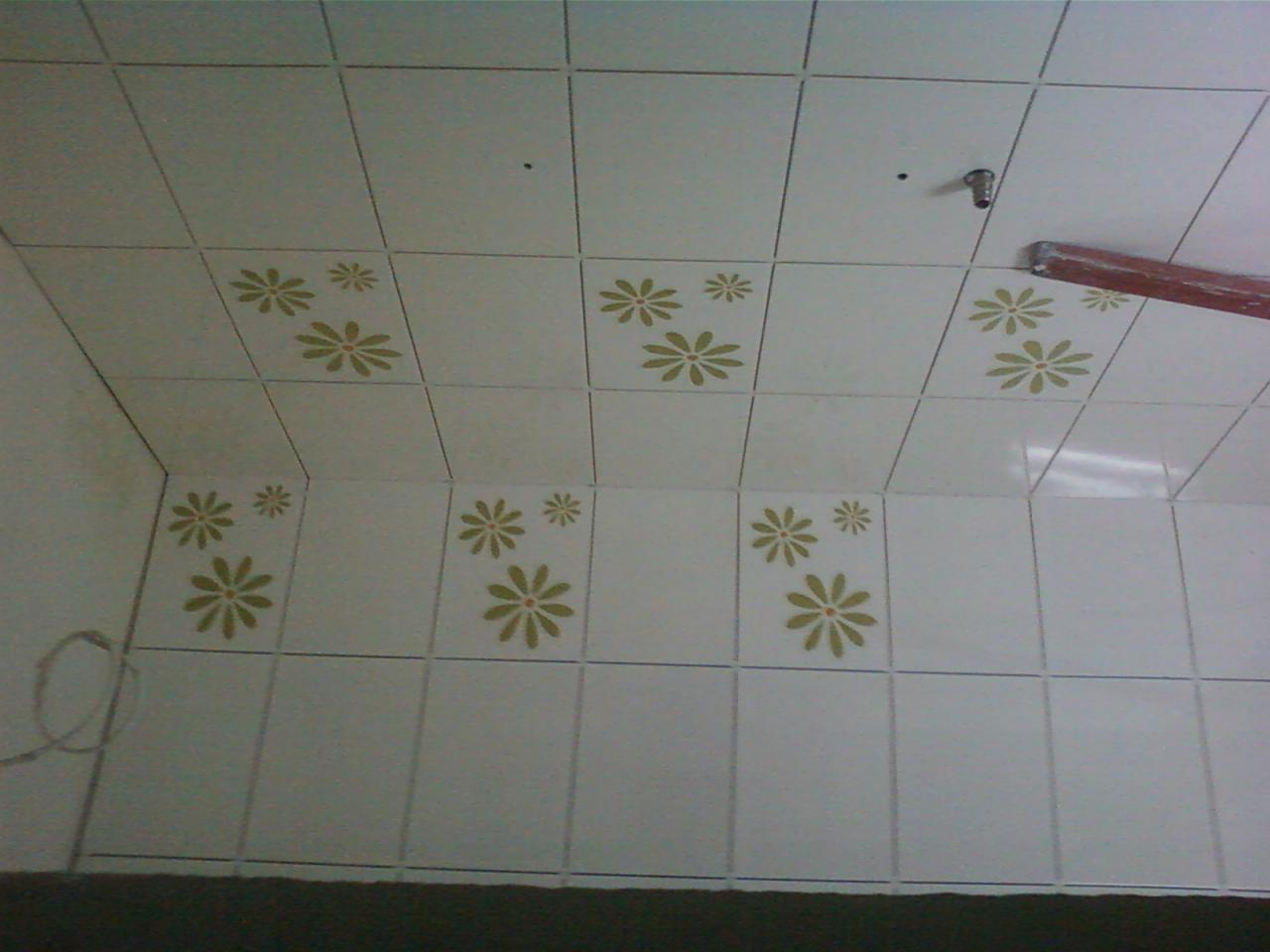 Białe, kwadratowe płytki ścienne i sufitowe z motywem kwiatowym w odcieniach oliwki, widoczne niedoskonałości fug i ślady po montażu armatury w rogu pomieszczenia.