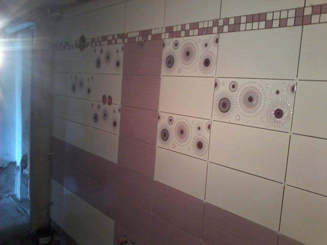 Ściana łazienki wyłożona jasnymi i różowymi płytkami z dekoracyjnym wzorem, widoczne niedokończone prace i surowy stan pomieszczenia w tle.