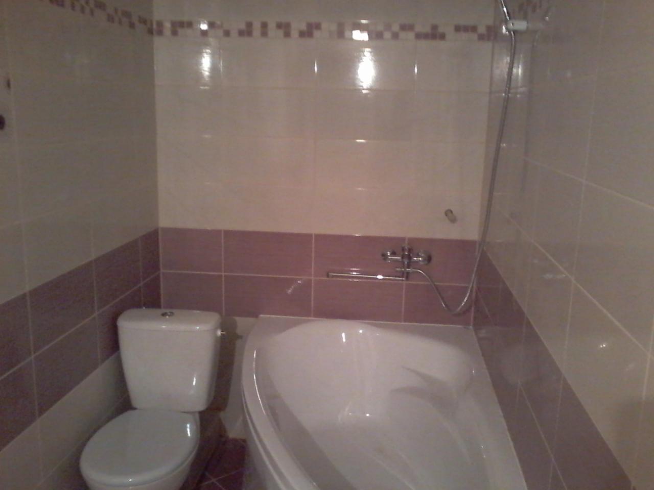 Wykończona łazienka z białą armaturą: toaleta i narożna wanna, ściany wyłożone jasnymi płytkami z bordowym pasem, chromowana bateria wannowa i słuchawka prysznicowa na ścianie.