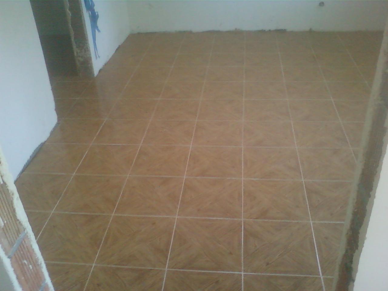 Ułożone, brązowe płytki podłogowe w kwadraty z jasnymi fugami w surowym pomieszczeniu, widoczne ślady po kleju na ścianach.