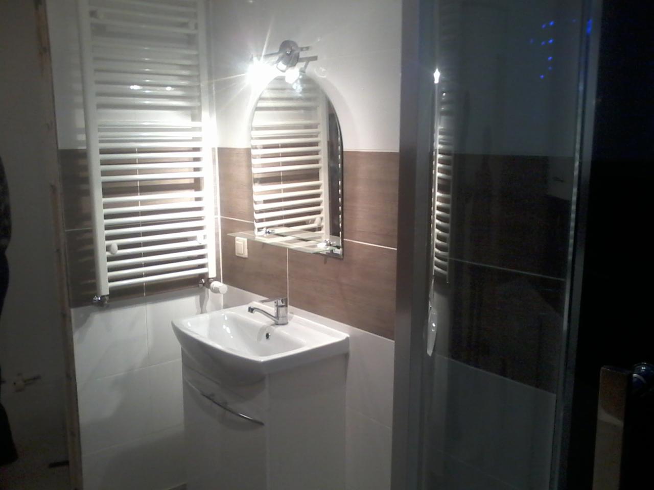 Nowoczesna łazienka z białymi płytkami, brązowym pasem dekoracyjnym, grzejnikiem drabinkowym, umywalką z szafką i kabiną prysznicową z ciemnego szkła.