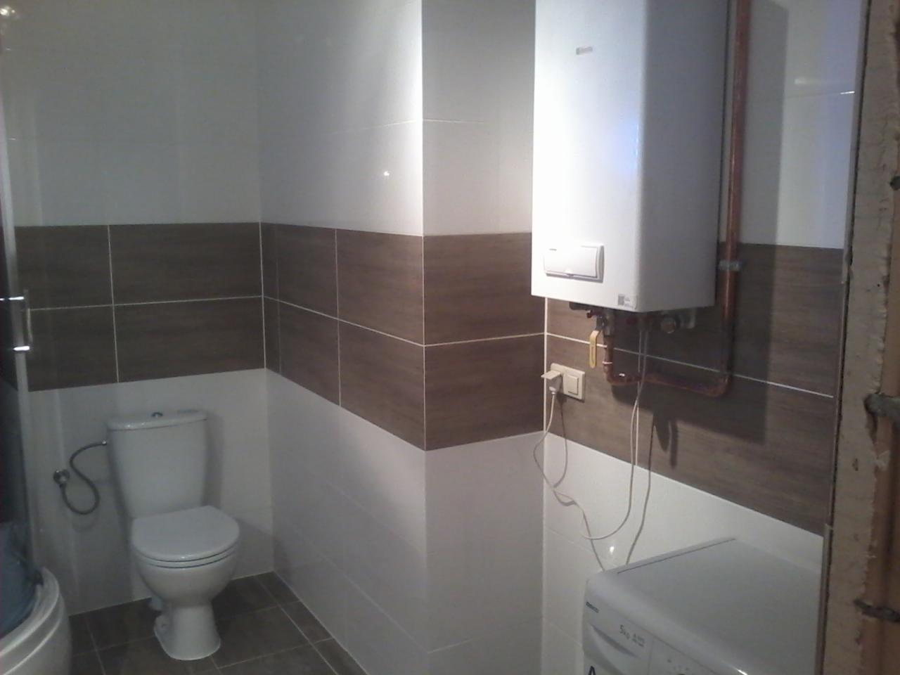 Wykończona łazienka z toaletą, pralką i piecem gazowym na tle białych i brązowych płytek, widoczne miedziane rury.