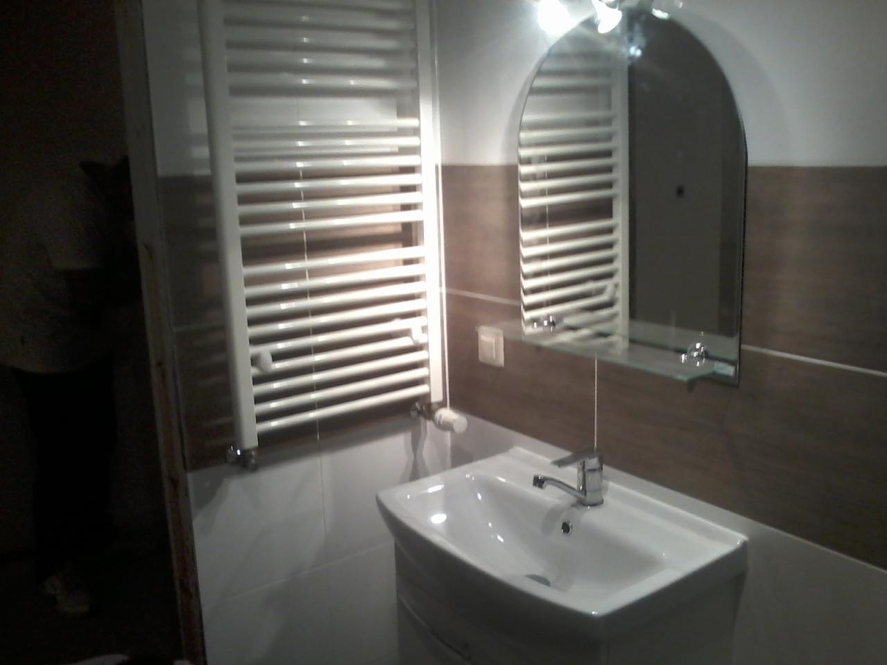 Biała umywalka z baterią, lustro w kształcie łuku i grzejnik drabinkowy na brązowo-białej ścianie w łazience, widoczne gniazdko elektryczne.