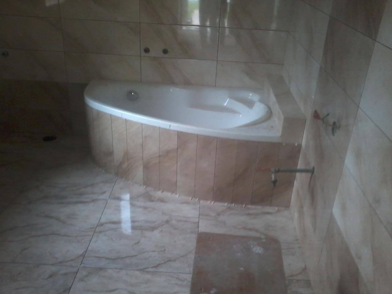 Narożna wanna z hydromasażem obudowana płytkami imitującymi marmur, dopasowane płytki na podłodze i ścianach, widoczne przyłącze wodne.