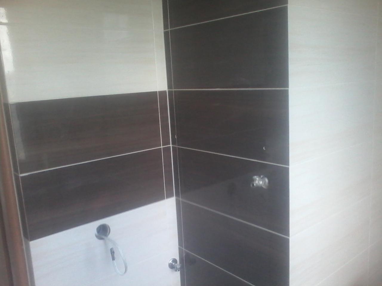 Ściana kabiny prysznicowej wyłożona jasnymi i ciemnymi płytkami w poziome pasy. Widoczne przyłącza wodne i odpływ.