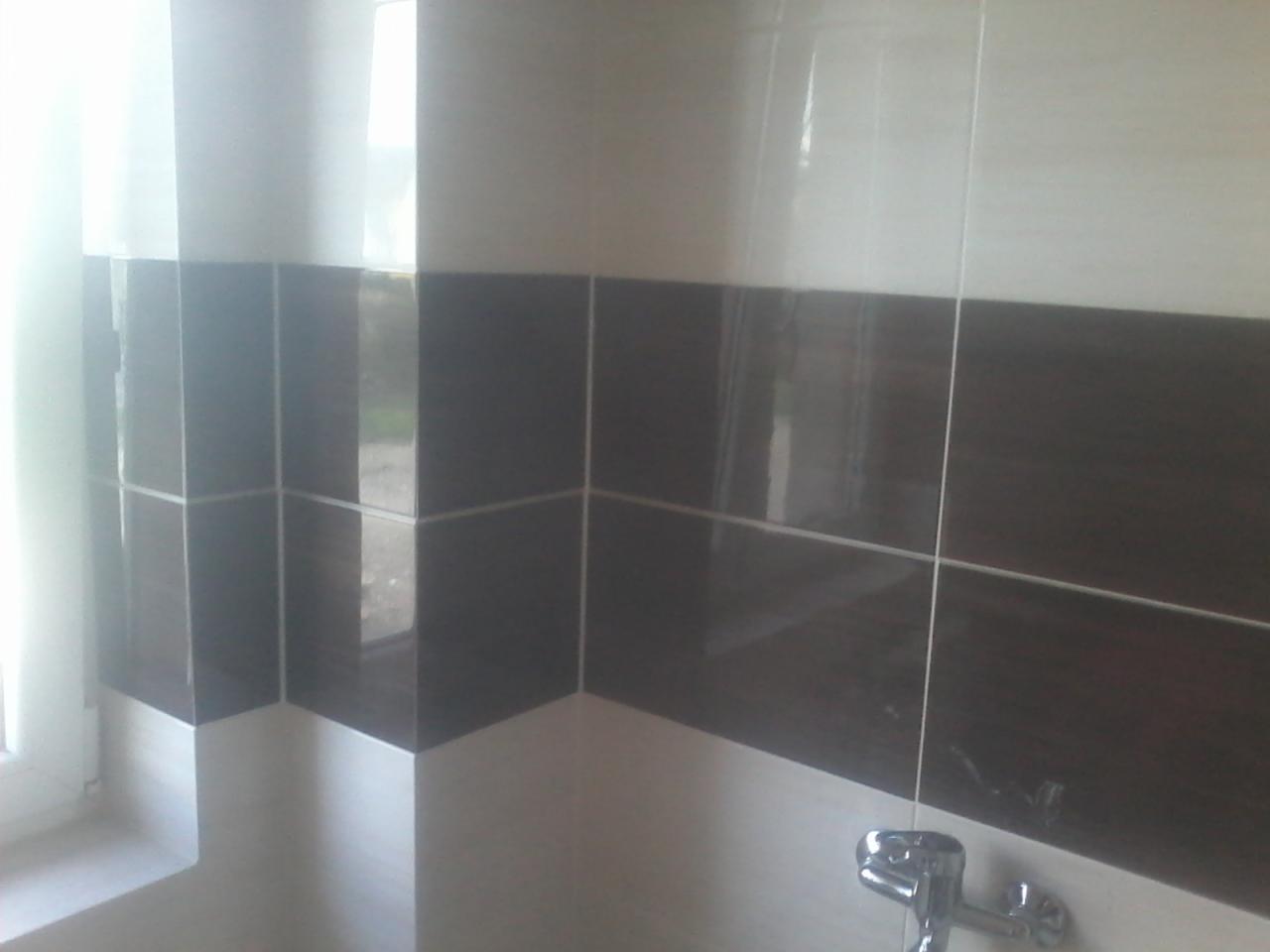 Fragment wykończonej ściany łazienki z dwukolorowymi, błyszczącymi płytkami ceramicznymi (białe i imitujące ciemne drewno) oraz widocznym fragmentem baterii umywalkowej.