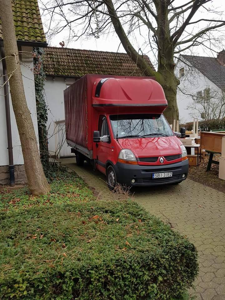 Czerwony samochód dostawczy Renault zaparkowany na brukowanej drodze przed domem z meblami w tle, prawdopodobnie podczas załadunku lub rozładunku.