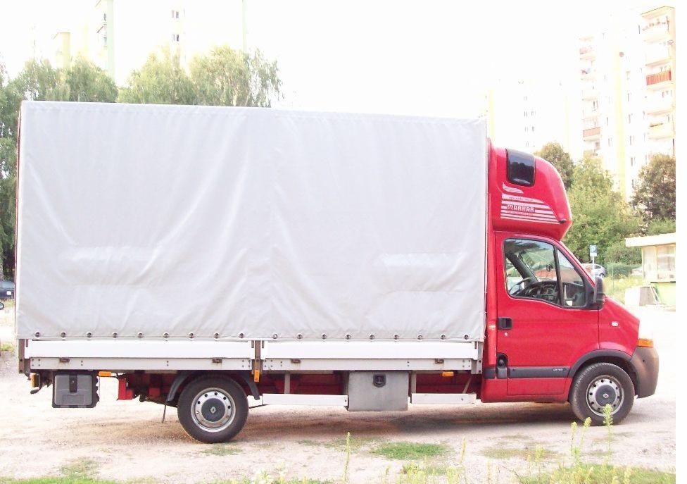 Czerwony samochód dostawczy z białą plandeką na tle bloku mieszkalnego i drzew.