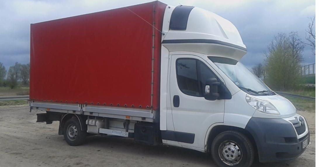 Biały samochód dostawczy z czerwoną plandeką, zaparkowany na poboczu drogi, pochmurne niebo w tle.