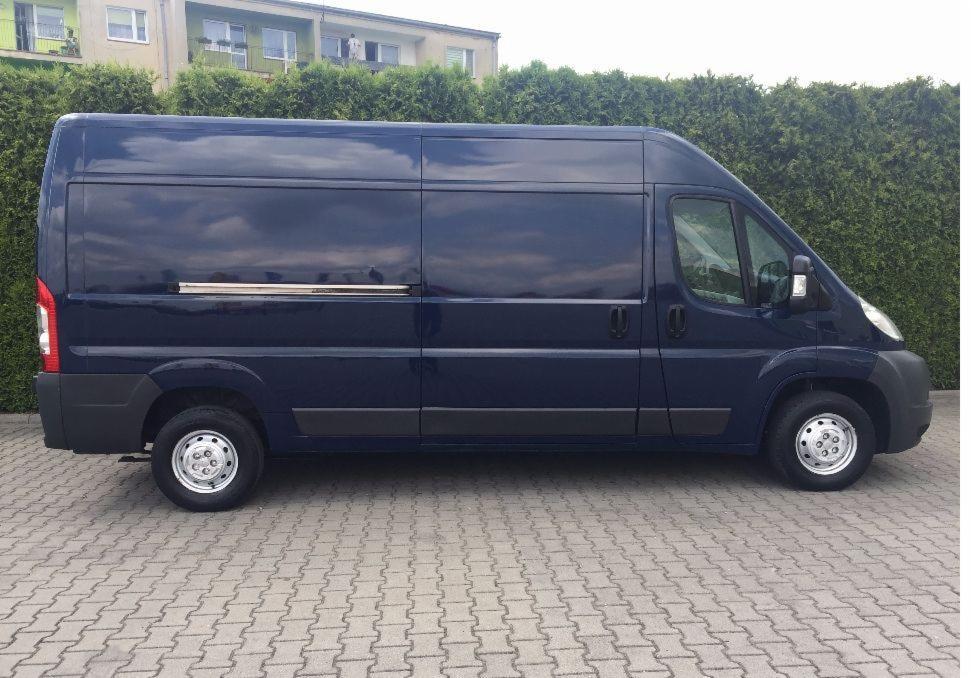 Niebieski samochód dostawczy z przesuwnymi drzwiami, zaparkowany na kostce brukowej, z odbiciem chmur na karoserii.