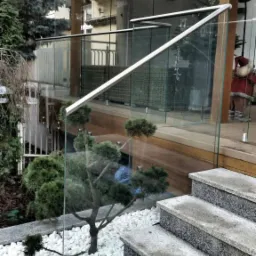 Szklana balustrada z metalową poręczą na tarasie z drewnianą podłogą, widok z dołu na schody betonowe i ogródek z białymi kamieniami oraz bonsai.