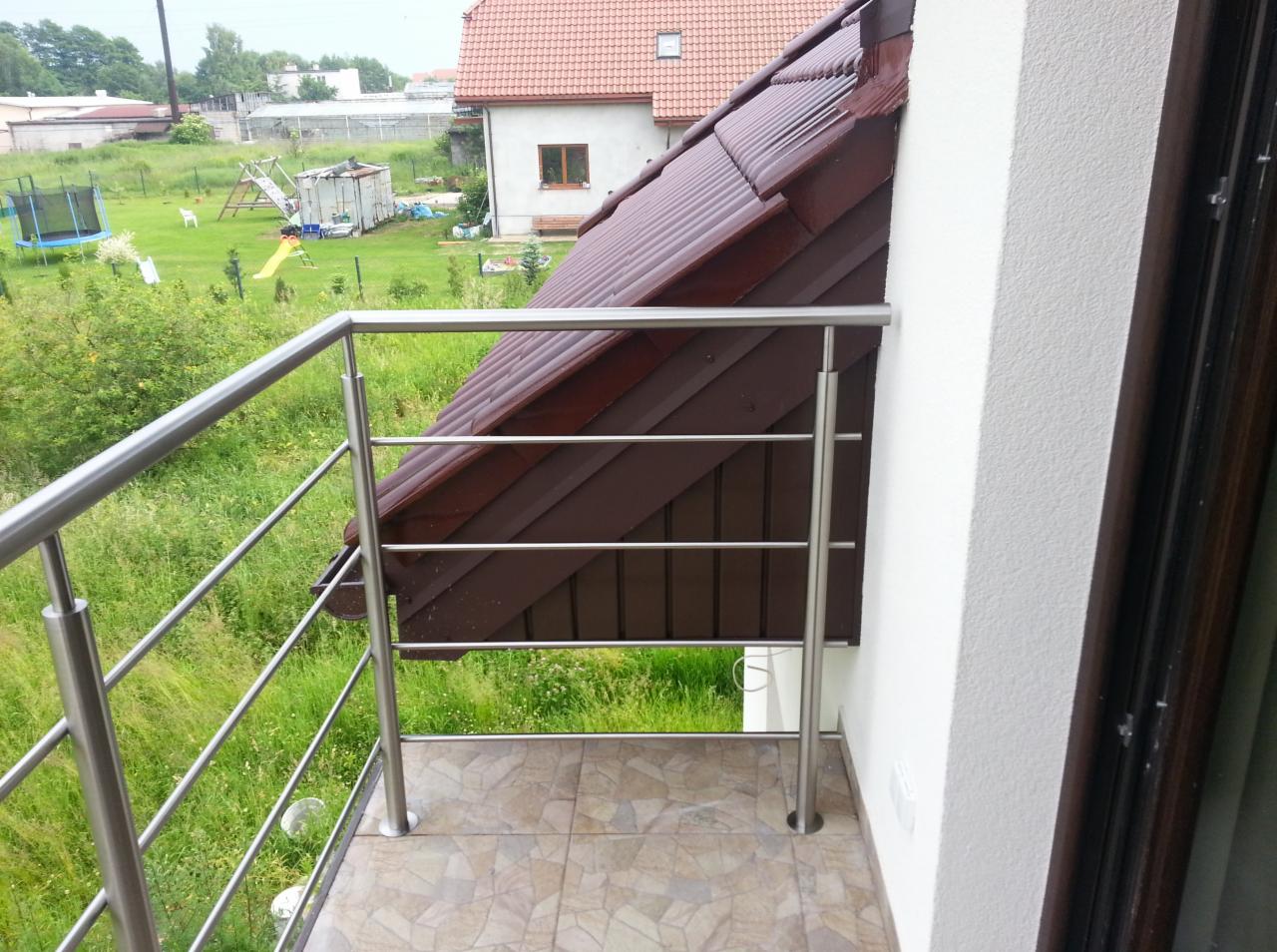 Balkon z nowoczesną, minimalistyczną balustradą ze stali nierdzewnej z czterema poprzeczkami, widok na zielony ogród z trampoliną i dom z brązowym dachem.