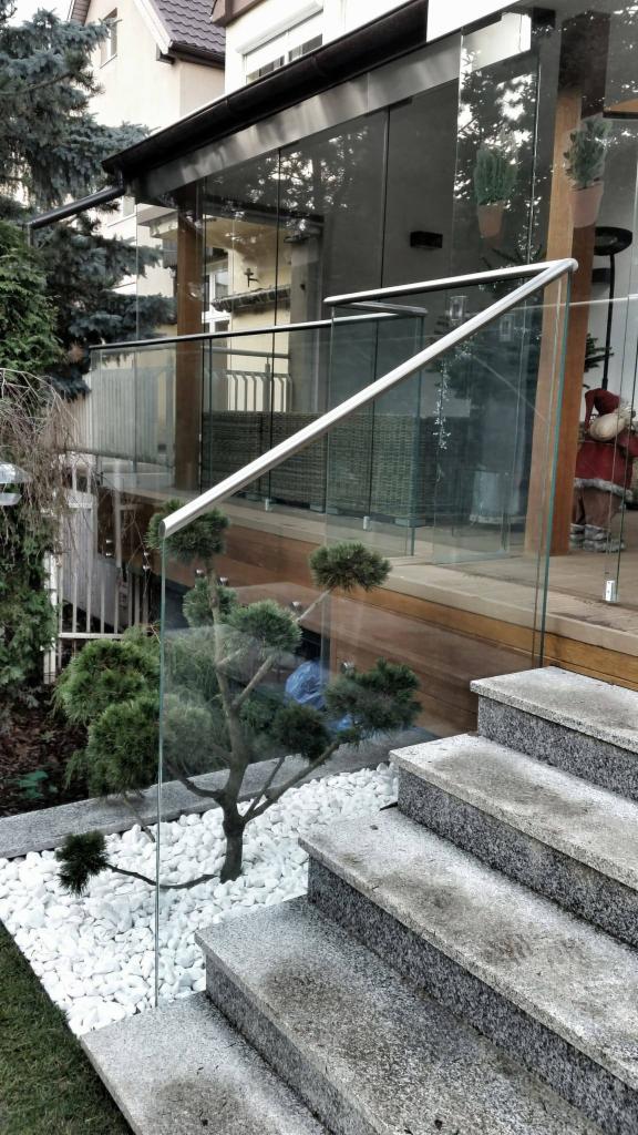 Szklana balustrada z metalową poręczą na tarasie z drewnianą podłogą, widok z dołu na schody betonowe i ogródek z białymi kamieniami oraz bonsai.