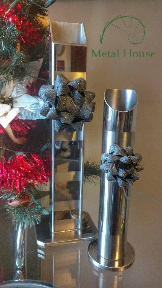 Dwa słupki z polerowanej stali nierdzewnej, ozdobione brokatowymi kokardkami, stoją obok choinki, widoczne logo firmy Metal House.