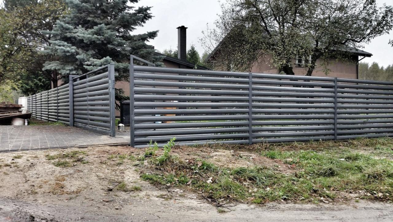 Nowoczesne ogrodzenie posesji z poziomych, metalowych profili w kolorze grafitowym, z widocznym fragmentem domu w tle.