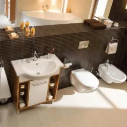 Nowoczesna łazienka z brązowymi płytkami, białą umywalką z szafką, toaletą i bidetem, uzupełniona o akcesoria łazienkowe i oświetlenie.